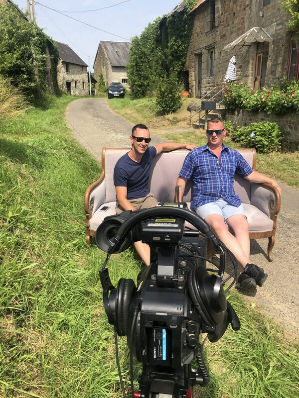 Yip and Paul sitting on a sofa in the lane through the hamlet, filmed by a television camera in the foreground